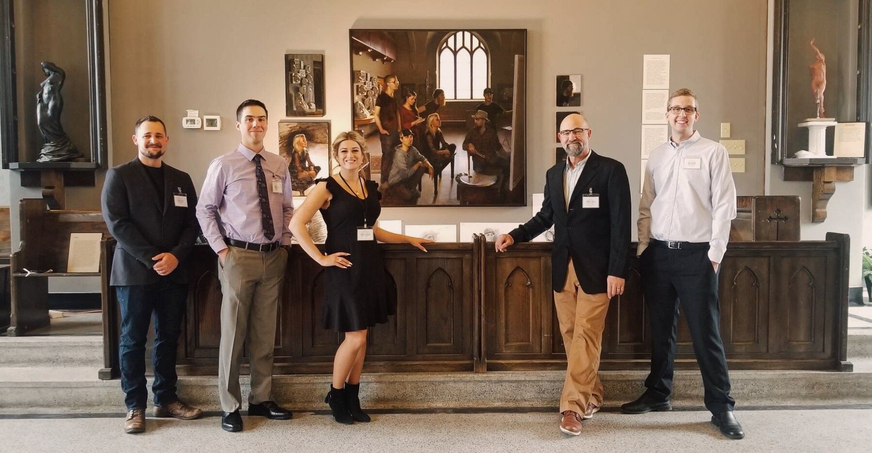 A group of five people, three men and two women, stand in a museum-like setting in front of a painting. They are dressed in business casual attire, and all are smiling. Various sculptures and artwork are displayed around them.