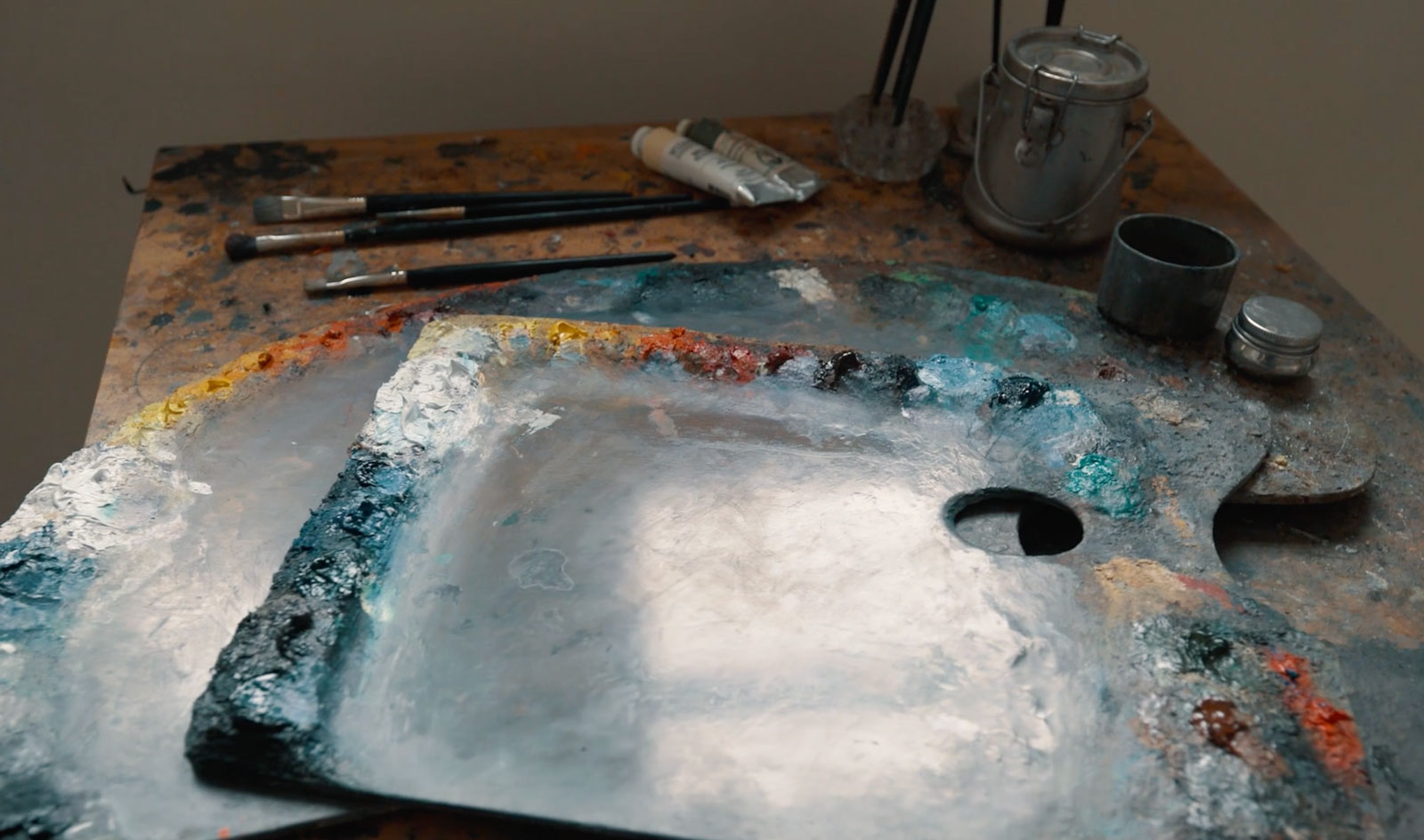 A close-up of an artist's workspace at a Duluth art school. Two paint palettes with mixed colors and used paintbrushes rest on a rustic wooden table. Nearby, a metal canister and other art supplies are visible, suggesting a creative setting.