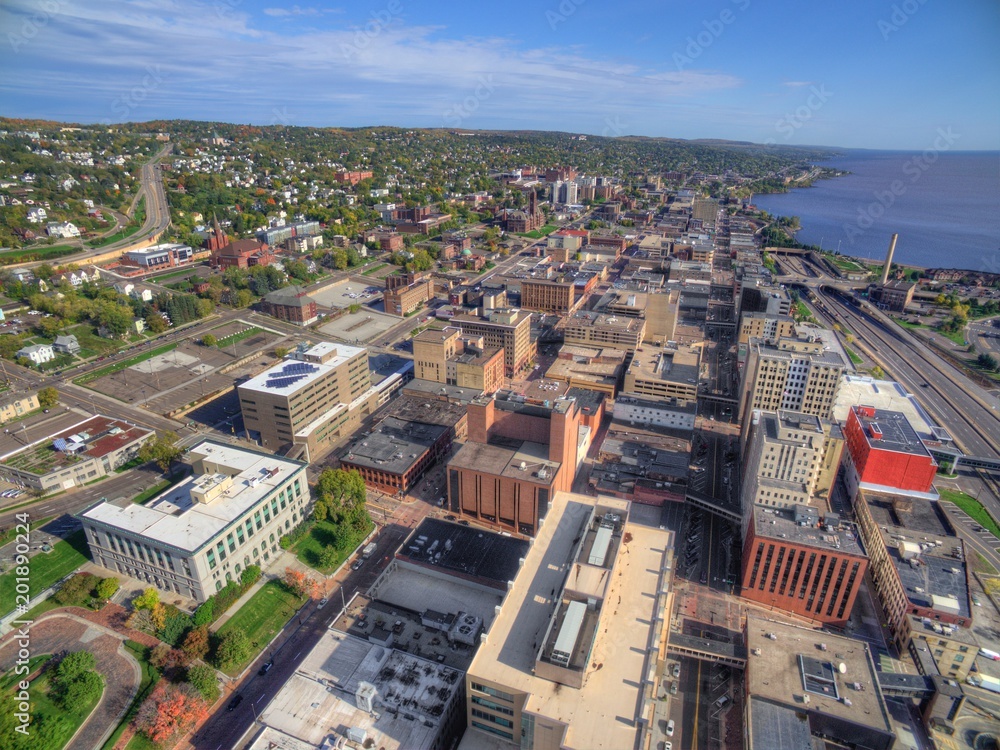 Aerial view of a city with a mix of modern and historic buildings. Streets are laid out in a grid pattern, with green spaces scattered throughout. A body of water is visible to the right, and the horizon shows a mix of city and countryside.