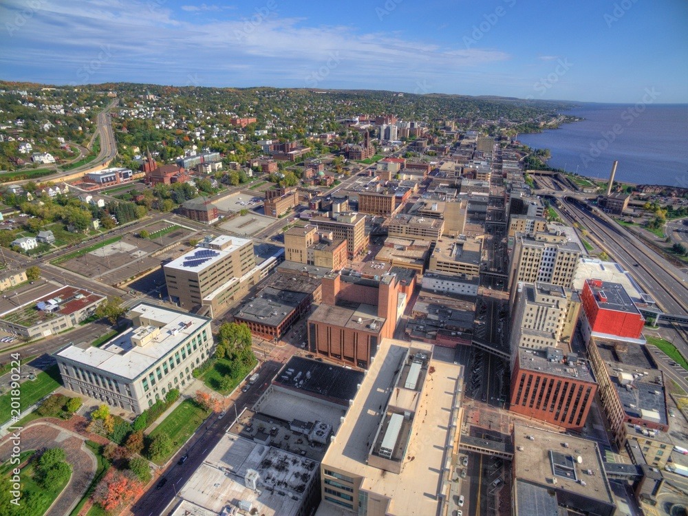 Aerial view of a cityscape near a large body of water. The city features a mix of modern and historic buildings, with tree-lined streets and a coastal road. Hills and greenery are visible in the background under a partly cloudy sky.