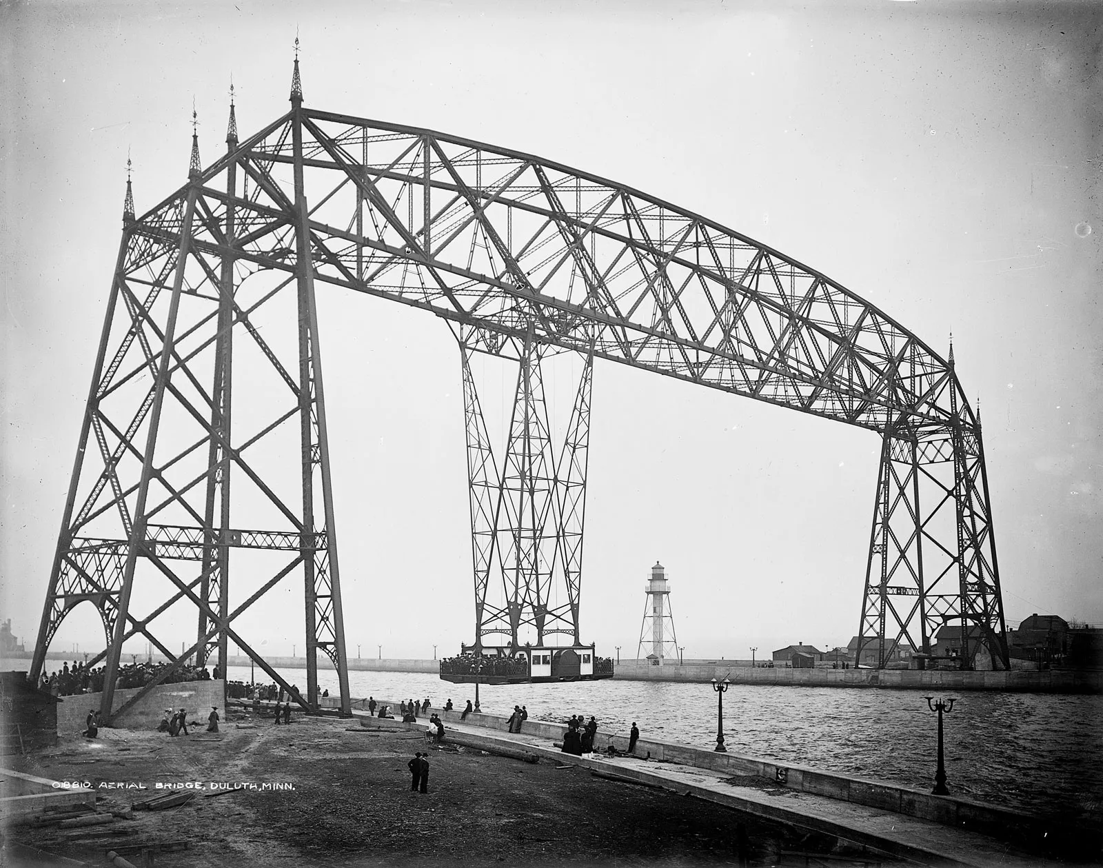 A large aerial lift bridge spans a body of water. People stand along the shoreline observing the structure. A lighthouse is visible in the background. The scene is in black and white, indicating an early 20th-century setting.