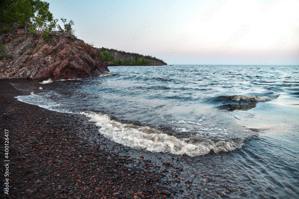 Rocky shoreline with small waves lapping against a pebble beach. A cliff covered with trees is visible in the background under a clear sky during sunset. The tranquil water reflects the soft hues of the evening light.