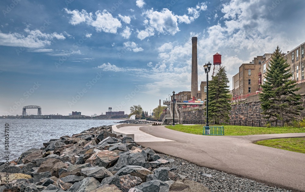 A scenic view of a waterfront with a path lined by trees and a stone wall. The background shows buildings, a bridge, and distant structures under a partly cloudy sky. The shoreline features large rocks.