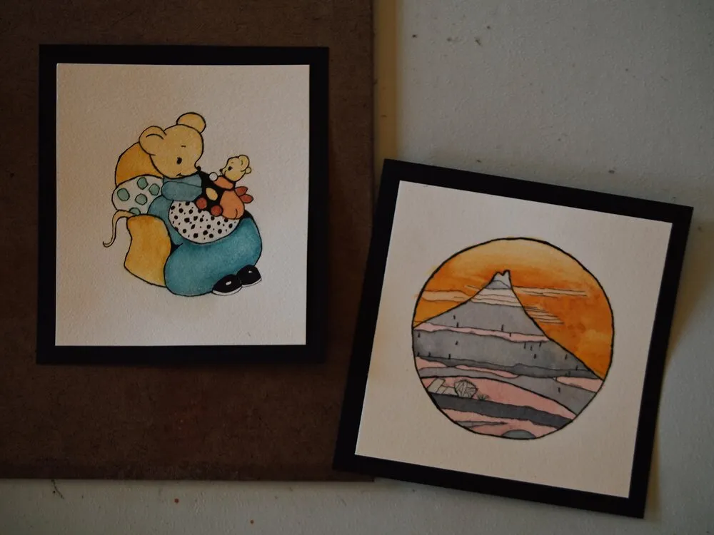 Two framed illustrations: one of a cartoon-style mouse sitting with a puppet on a couch, and the other of a mountain landscape at sunset with an orange sky, presented in a circular frame. Both images are placed against a neutral background.