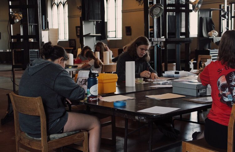 People sit at tables in a dimly lit room, engaged in various arts and crafts activities. The room features large windows and wooden chairs. Supplies and water bottles are scattered around the tables.