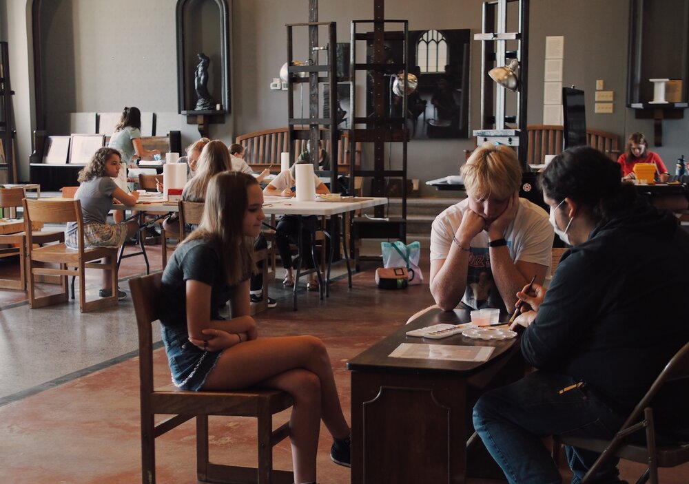 A group of people engaged in various activities in a spacious room with high ceilings. Some are seated at tables, working or chatting, while others are walking around. The room has a mix of modern and classical decor elements.