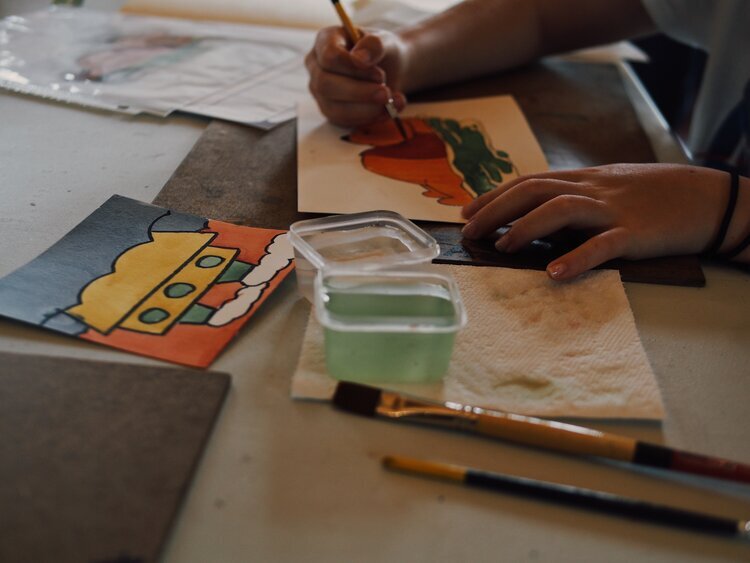 A person paints colorful designs on paper at a table. One painting depicts a vibrant character with a yellow hat and red attire. Art supplies, including brushes and paint containers, are scattered around.