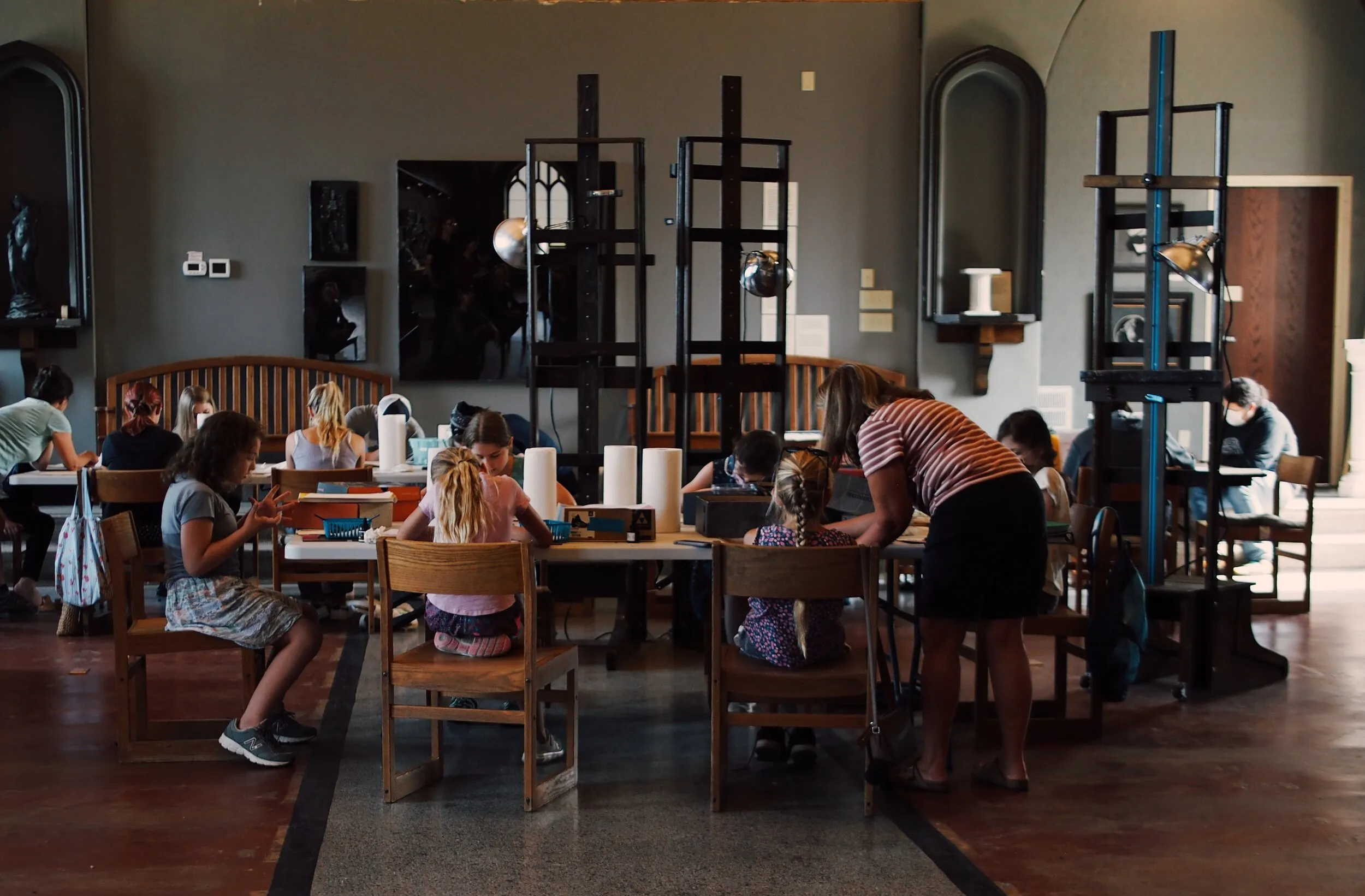 A group of children sitting at tables in an art classroom, working on projects. An adult is assisting them. The room is spacious with various art tools and equipment visible, and large windows provide natural light.