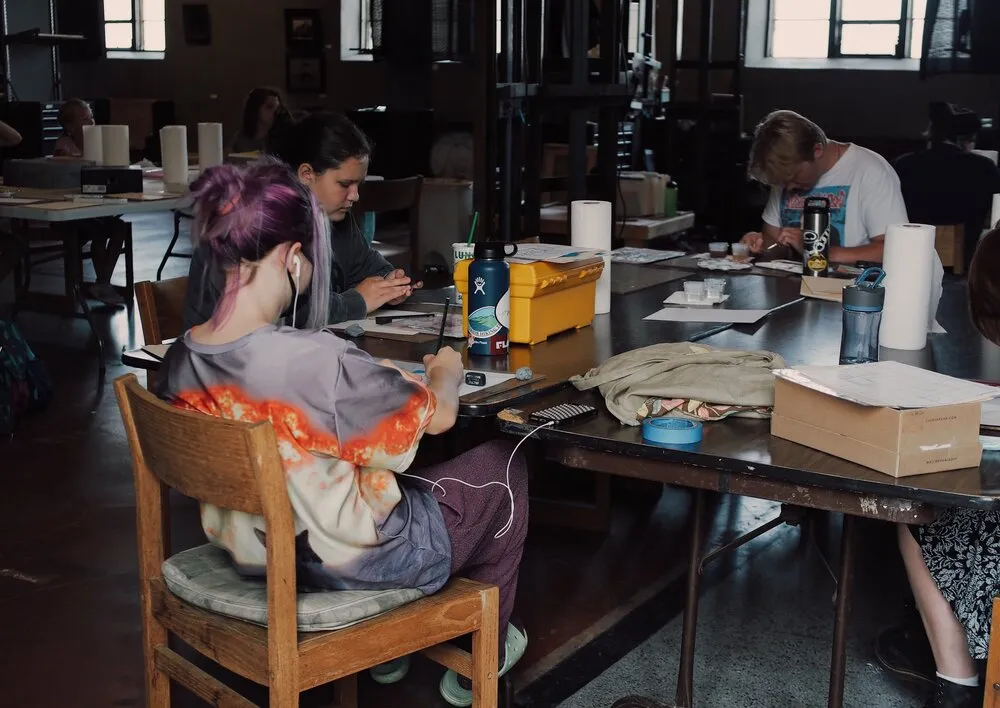 People sitting at a large table in a workshop setting, focused on various art and craft projects. The room has a casual atmosphere with supplies visible, and large windows providing natural light.