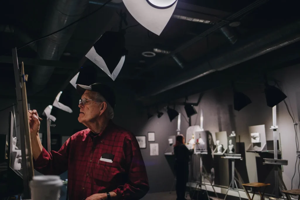 An older man in a red plaid shirt and cap is painting in a studio filled with easels and spotlights. The dark room is lit by overhead lights, and another person is visible painting in the background.