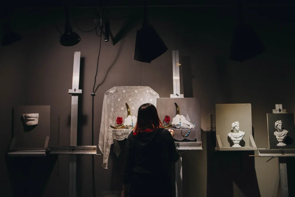 A person is painting in a dimly lit art studio. The workspace has sculptures and paintings of classical busts and lips displayed on easels. Red roses and draped fabric add decorative elements to the scene.