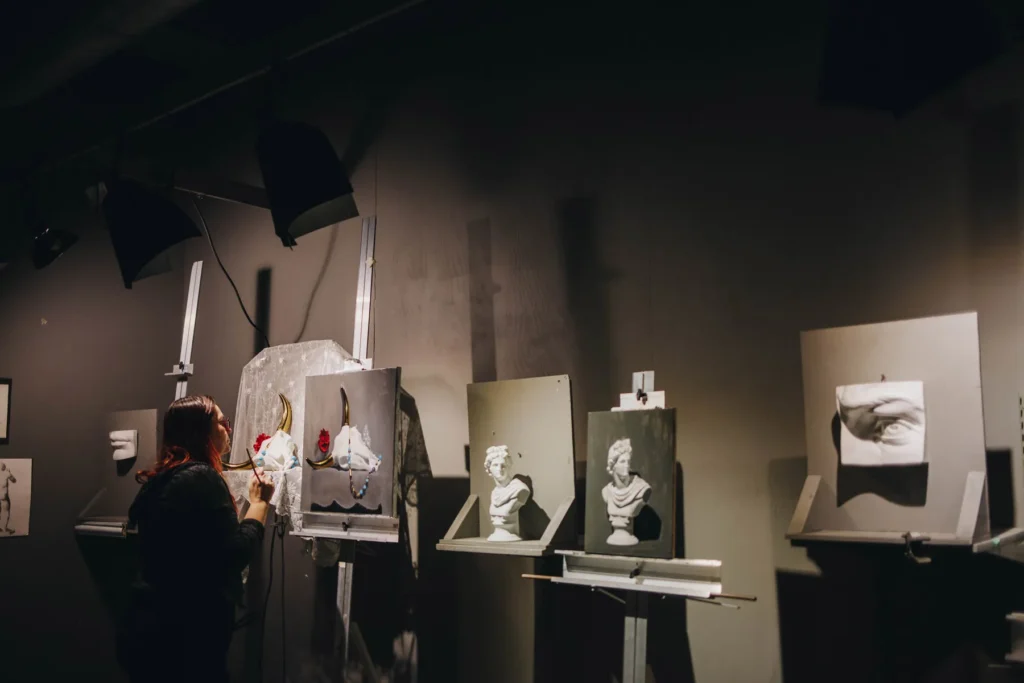 A dimly lit art studio with an artist painting on a canvas. Several plaster sculptures of facial features and busts are mounted on easels nearby. The room has a creative and moody atmosphere.