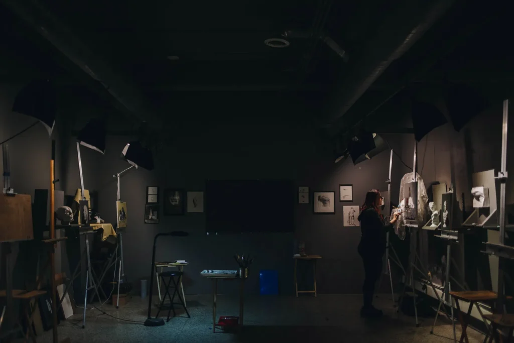 A dimly lit art studio with a person standing, working on a sculpture. Various artworks are displayed on the walls, and several spotlights illuminate the workspace. Art supplies and tools are scattered around the room.