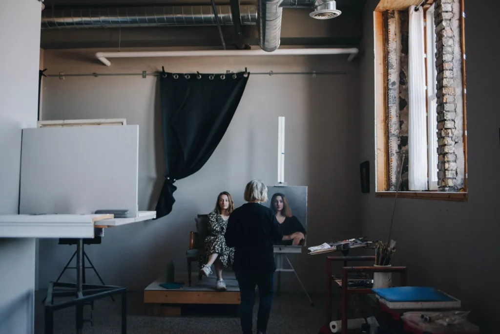 An artist paints a portrait of a woman sitting on a platform in an art studio. The studio has large windows, an easel, and various art supplies scattered around. A dark curtain hangs on the wall behind the platform.