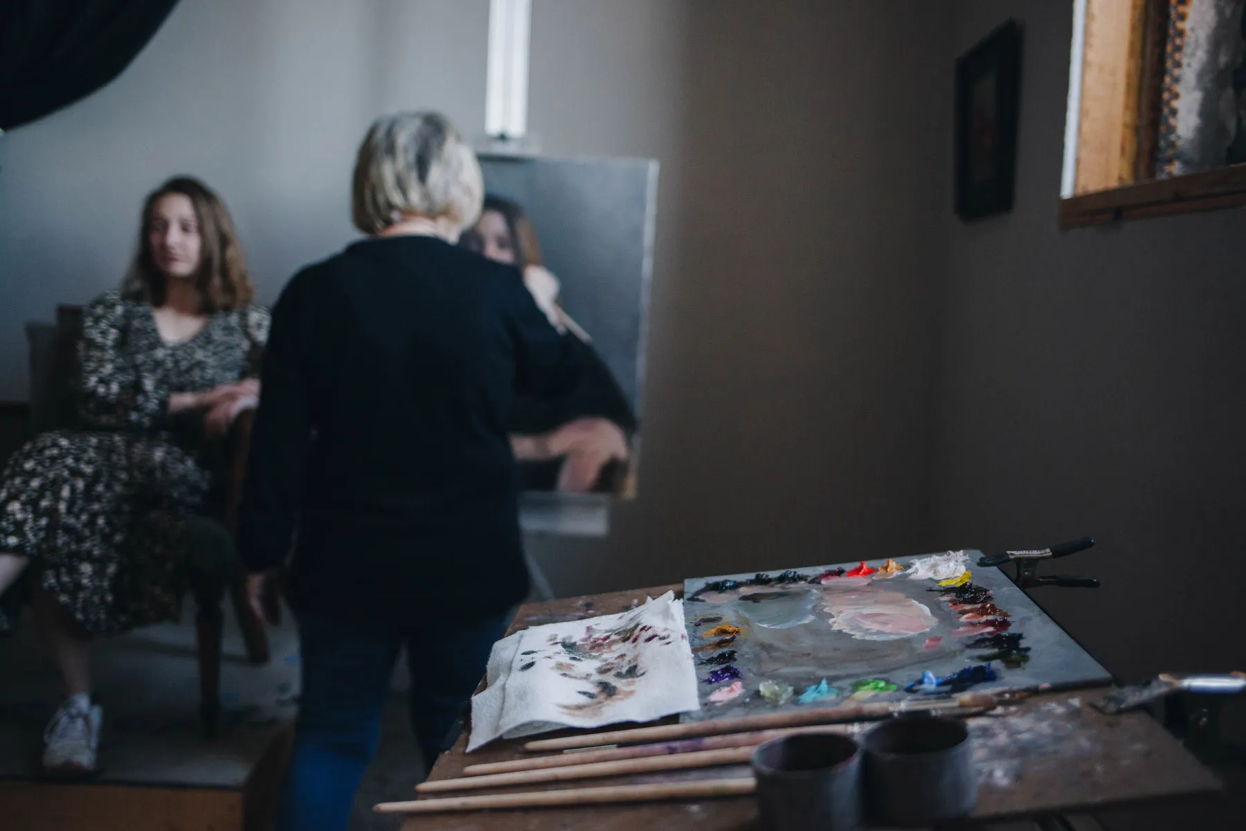 A woman with short hair stands at an easel, painting a portrait. The canvas shows a woman sitting in a chair in the background. A palette with various colors and brushes is in the foreground. The room is softly lit.