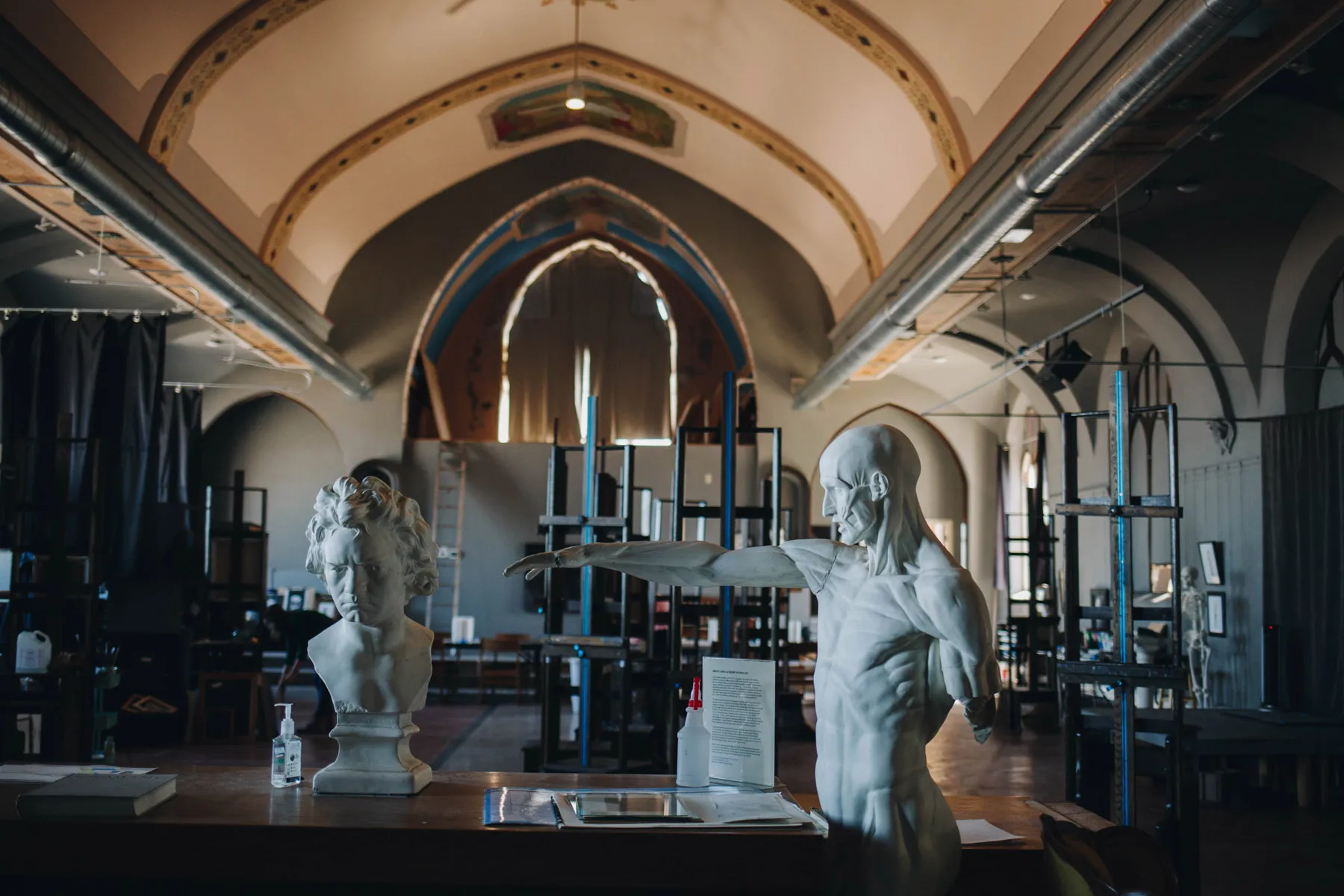 An art studio with a high arched ceiling features two sculptures: a bust of a woman and an anatomically detailed male figure with an outstretched arm. Easels and art supplies are visible in the background.