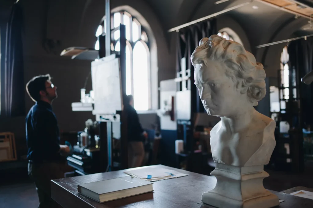 A marble bust of Beethoven sits on a wooden table in a room with arched windows. Two people are blurred in the background, focusing on art-related activities. The room is lit by natural light from the windows.