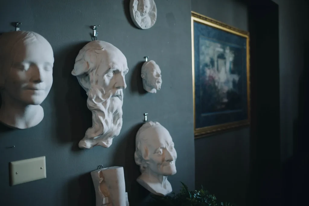 A dimly lit room with a dark wall displaying several white plaster busts of faces in profile and full view. A framed painting with a golden border hangs in the background, and a small plant is partially visible at the bottom.