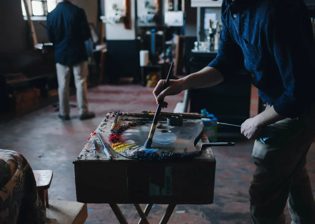An artist in a dimly lit studio paints on a palette with a range of colors. Another person stands in the background. The room is filled with art supplies and creative clutter, creating an atmosphere of artistic focus and activity.