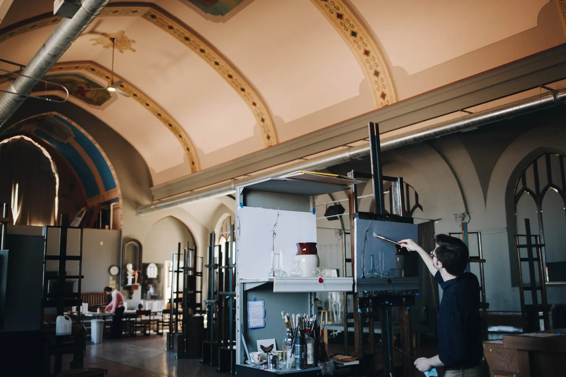 A person paints on a canvas in a spacious art studio with high ceilings and arched windows. The studio is filled with art supplies and easels, and another person is seated in the background.