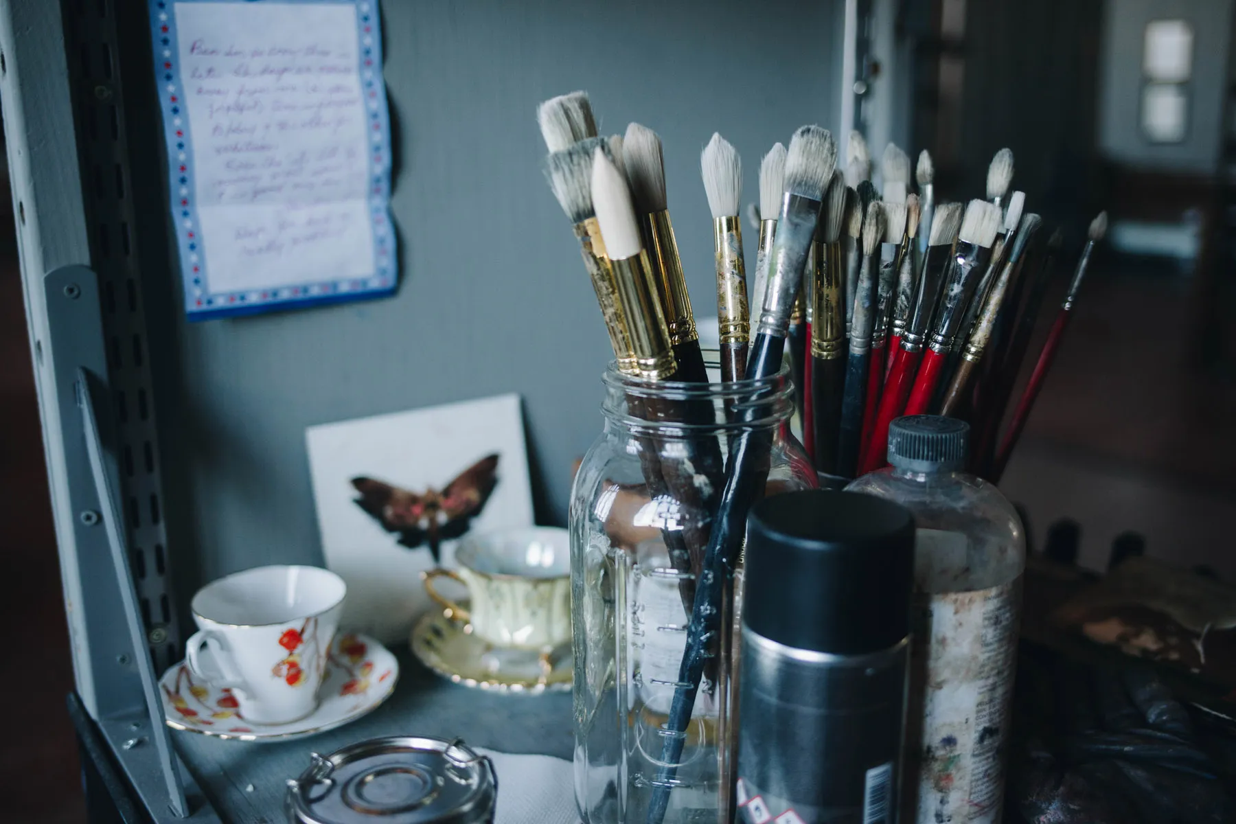 A collection of paintbrushes in a glass jar, surrounded by art supplies on a shelf. Nearby are teacups, a saucer, a spray can, and a small butterfly illustration. A handwritten note is pinned on the back wall.