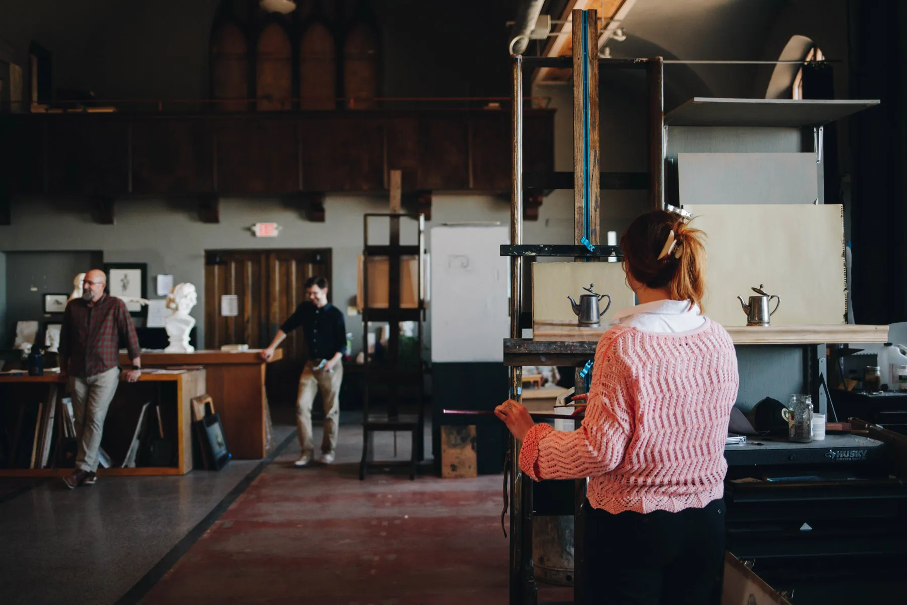 A woman in a pink sweater stands in an art studio, observing two men near a door. The studio is filled with art supplies, sculptures, and a warm ambiance with rich wooden tones and large windows.
