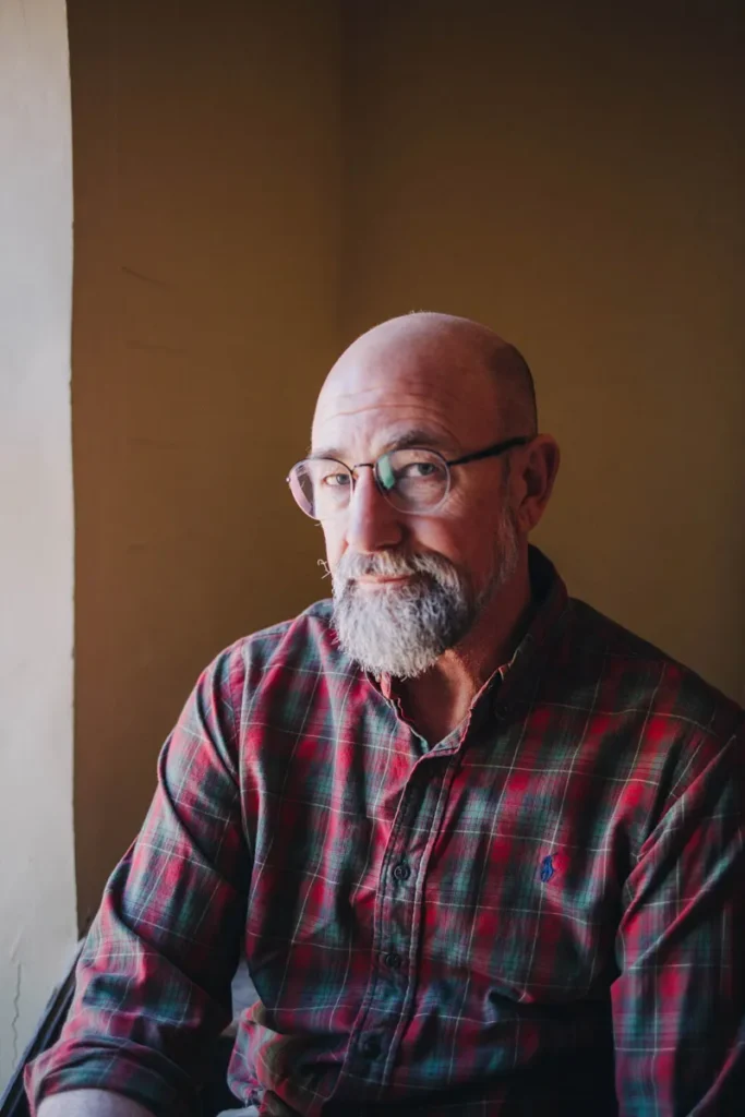 A bald man with a gray beard and glasses sits by a window, wearing a red and green plaid shirt, in a softly lit room.