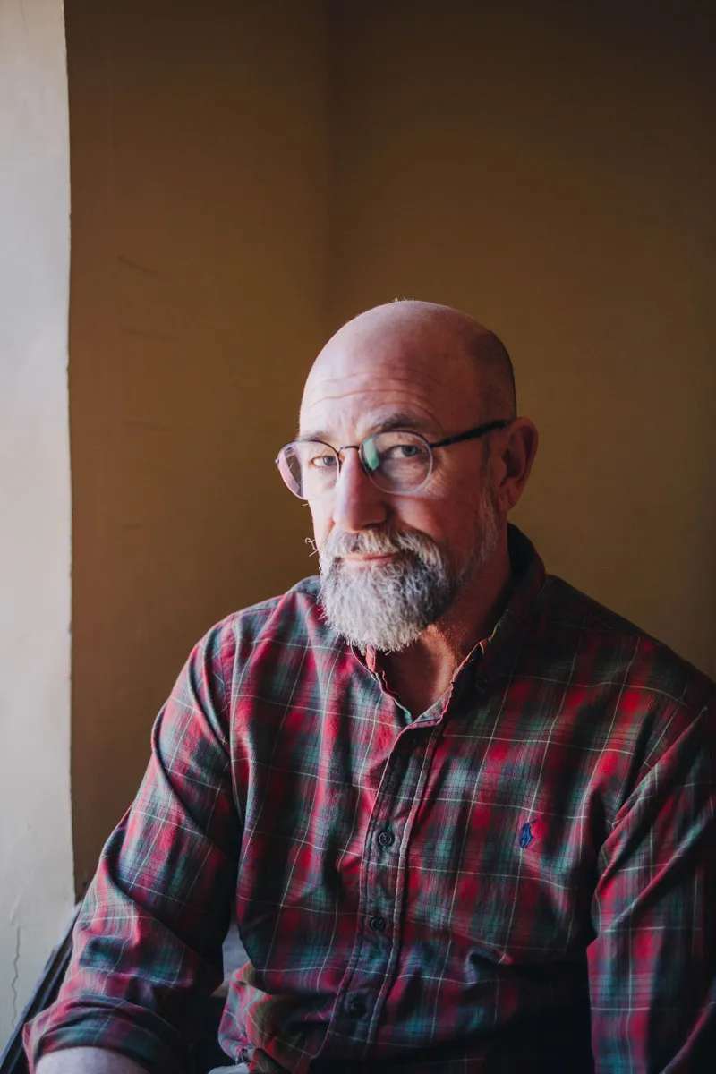 A bald man with a gray beard and glasses sits by a window, wearing a red and green plaid shirt, in a softly lit room.
