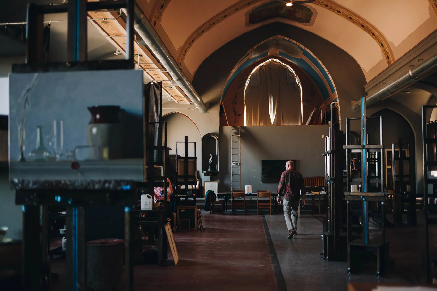 A person walks through an art studio with high arched ceilings and large windows. The room is filled with easels and art supplies, creating an inspiring workspace. Warm lighting adds a tranquil atmosphere.