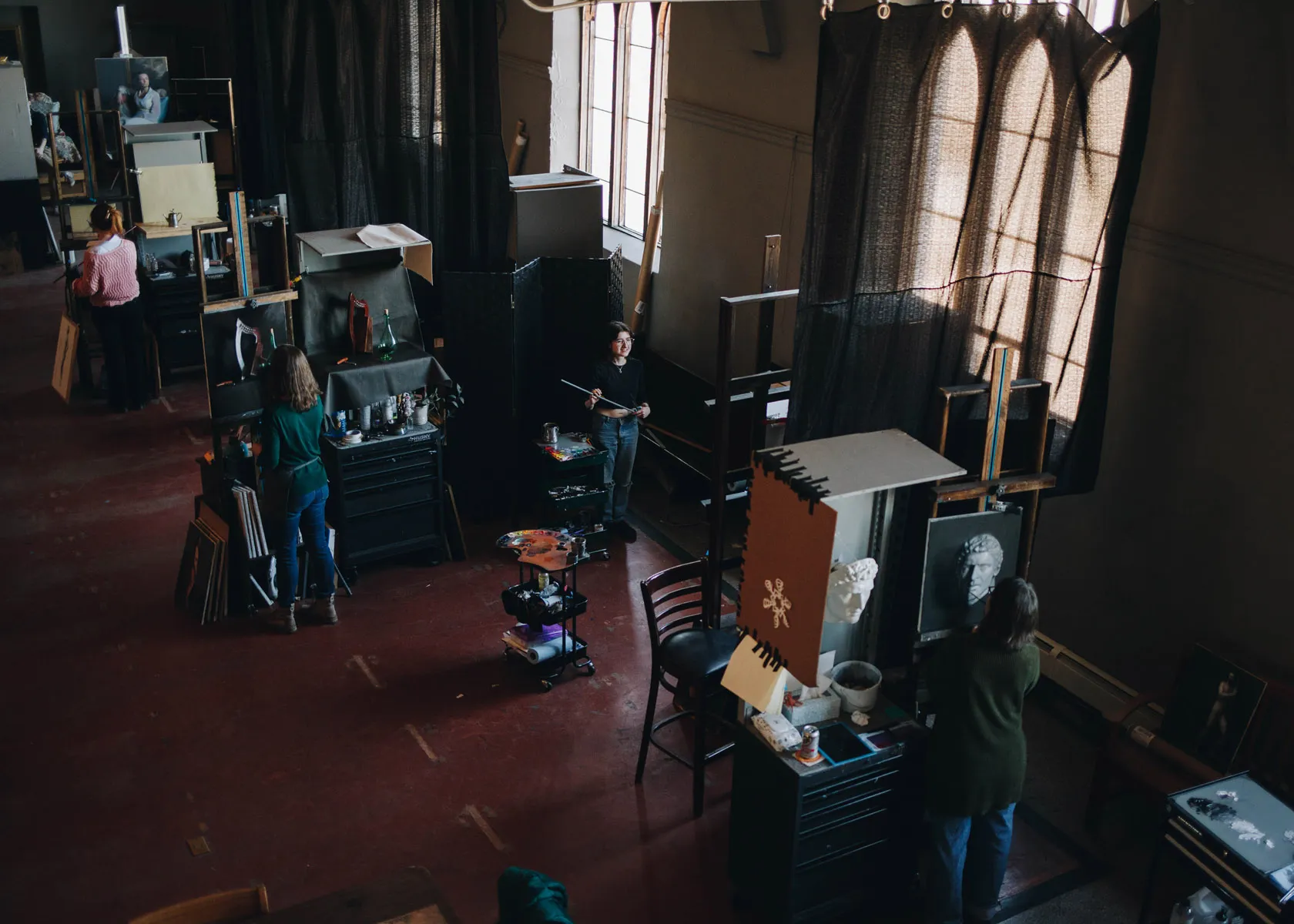 A spacious art studio with several people working at individual stations. Easels, paintings, and art supplies are visible. Large windows allow natural light to illuminate the space. The floor is red, and black curtains add contrast.