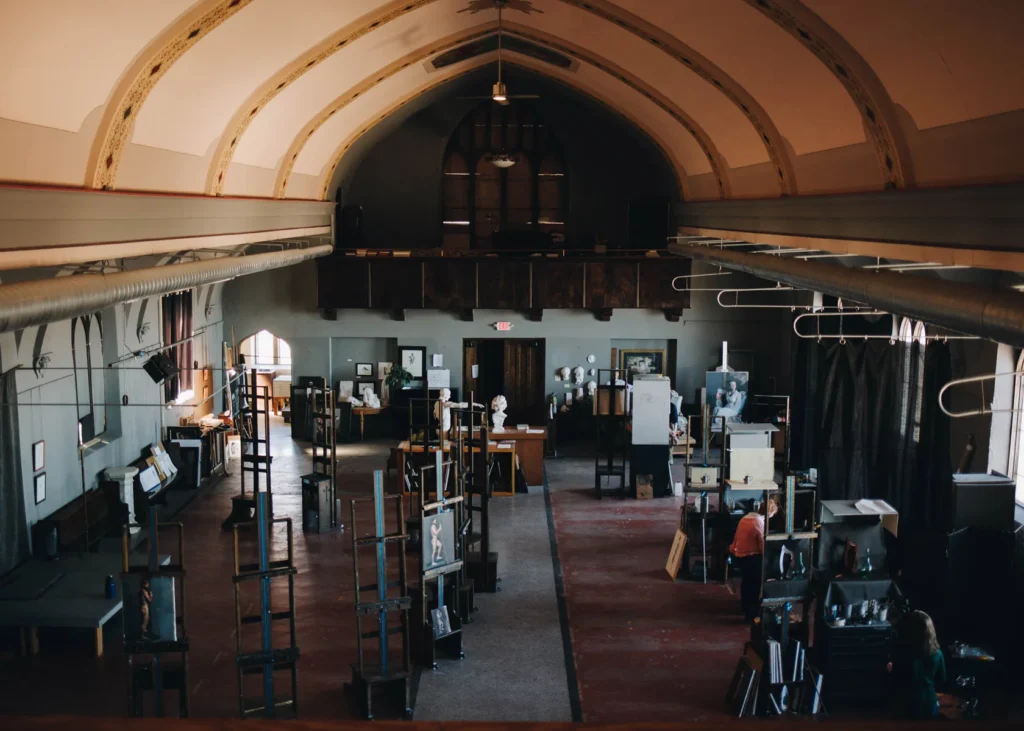 A spacious art gallery inside a repurposed church with a high vaulted ceiling. Various artworks and sculptures are displayed on stands throughout the room. Two people are observing the exhibits. Natural light filters through arched windows.