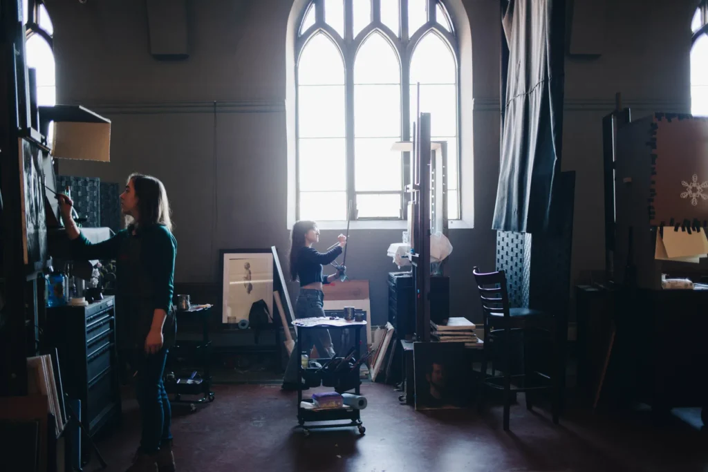 Two artists work in a dimly lit studio with high arched windows. One paints on an easel, while the other arranges materials. Art supplies and framed artworks are scattered around the room, creating a creative and focused atmosphere.