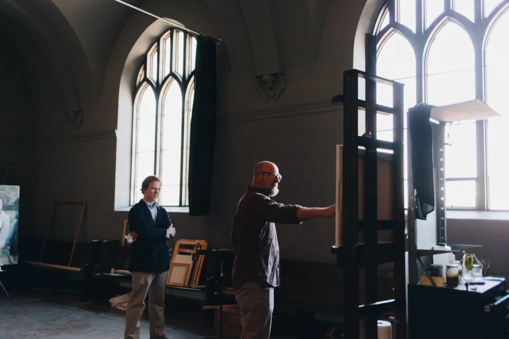 Two people in an artist's studio with arched windows. One is painting on a large canvas, while the other observes. The room is dimly lit with art supplies and framed canvases scattered around.
