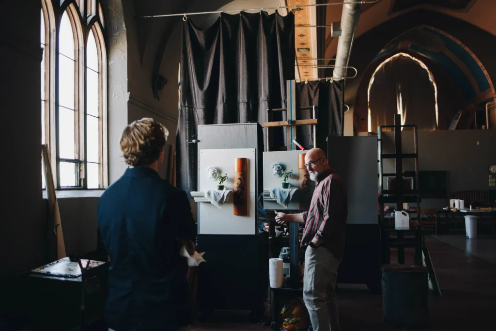 Two people in an art studio observe two abstract paintings on easels. The room is dimly lit with high ceilings, arched windows, and various art supplies around. One person gestures towards the paintings.