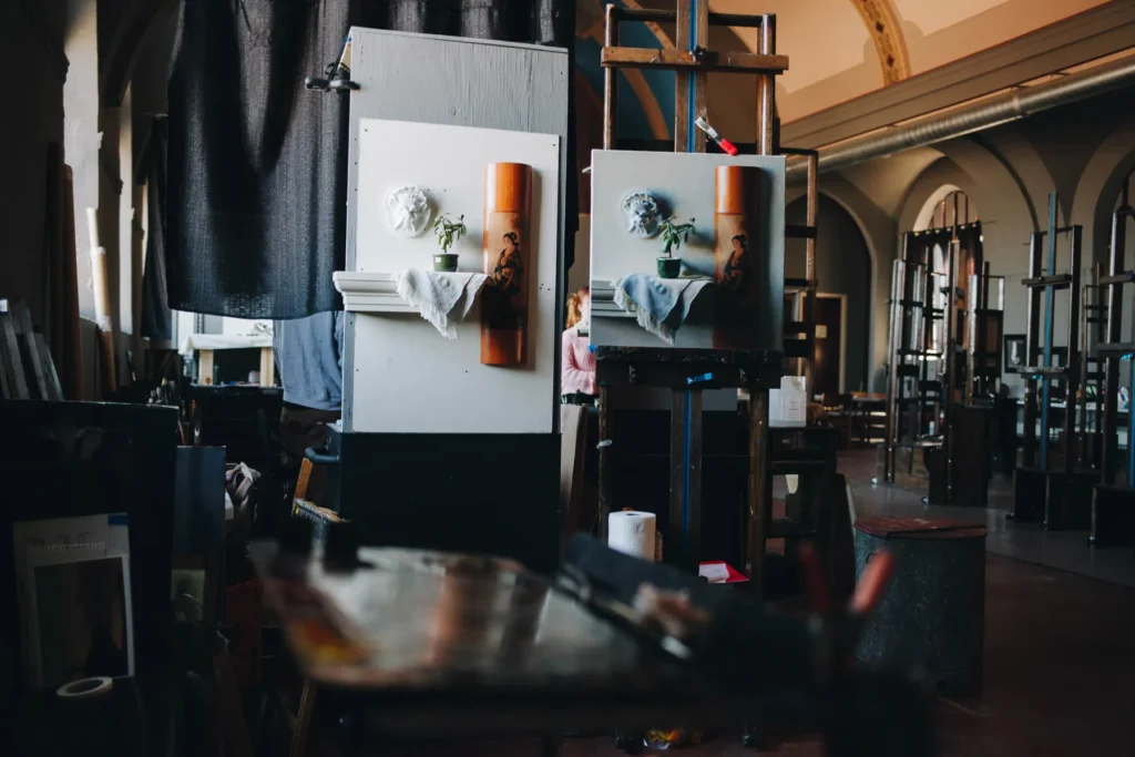 An artist's studio with a still life painting on an easel. The painting depicts a white sculpture, a vase with flowers, and a cloth on a wooden shelf. The dimly lit room has high ceilings and various art supplies scattered around.