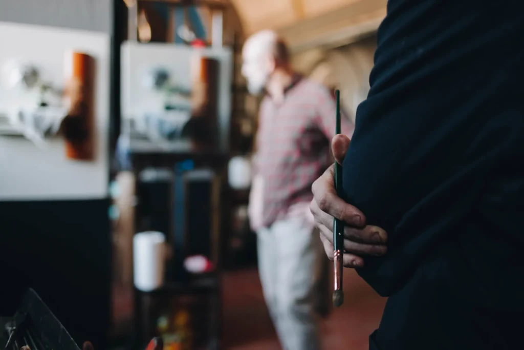 A person holds a paintbrush in the foreground, with their arm crossed. In the background, a man in a checkered shirt stands in a slightly blurred art studio, surrounded by canvases and art supplies. The scene conveys a creative atmosphere.