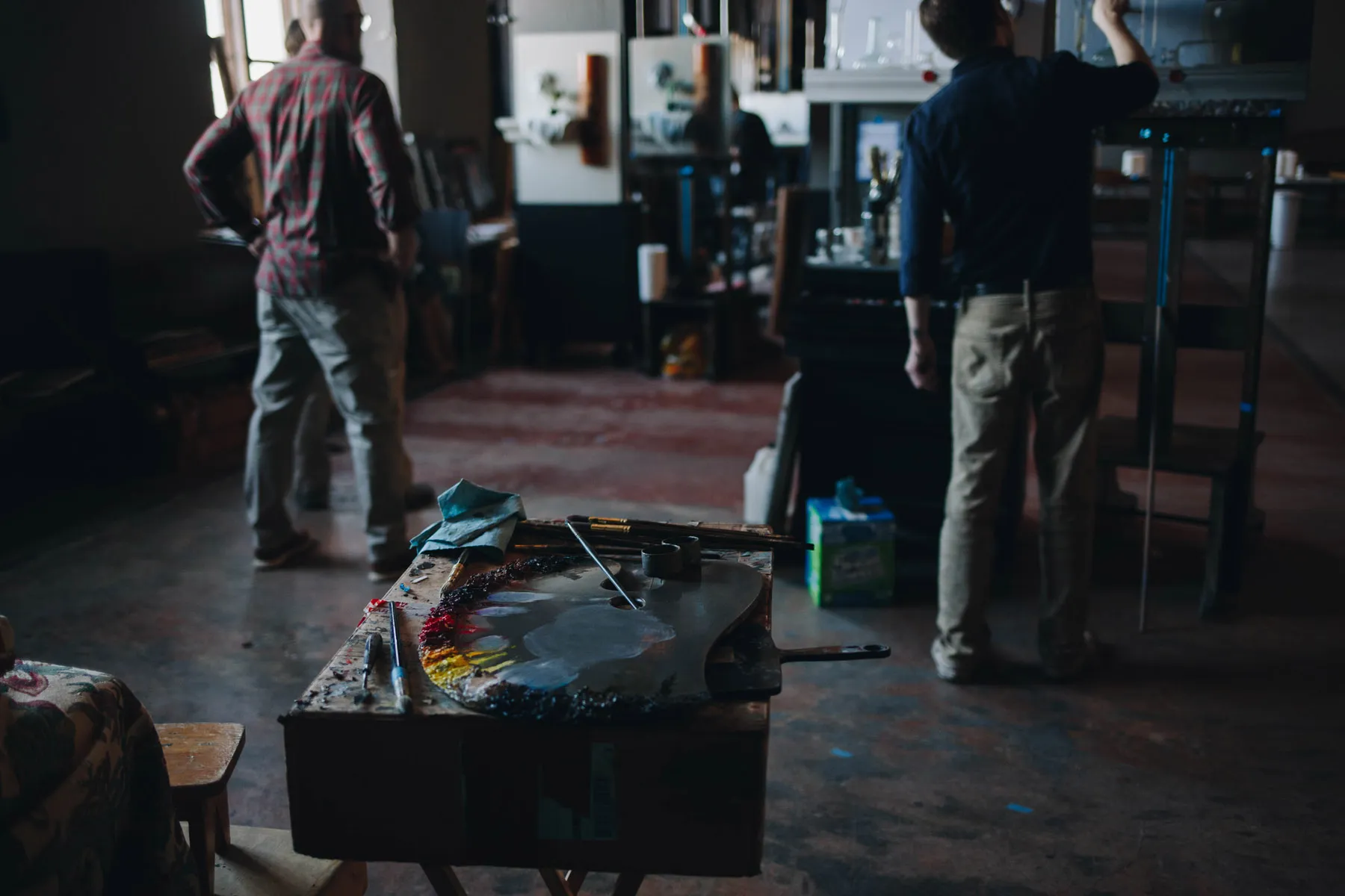 Two people are in an art studio. One is painting on a canvas placed on an easel, while the other stands nearby. In the foreground, a wooden table holds palettes and brushes, with soft light illuminating the space.