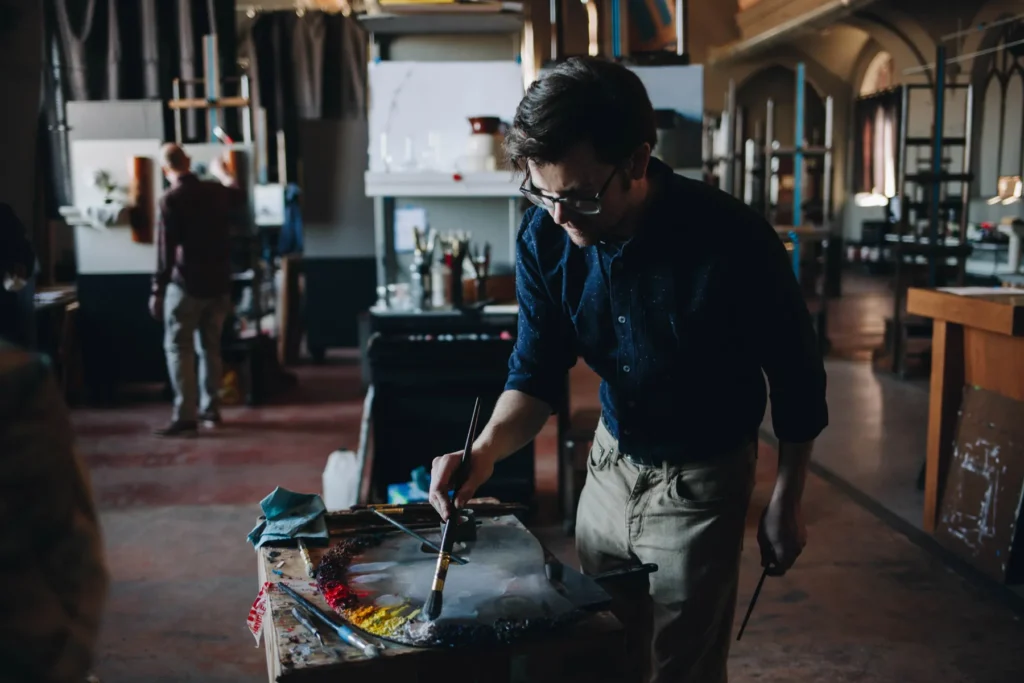 A person painting on a canvas in a dimly lit art studio, wearing glasses and a dark shirt. The studio is filled with art supplies and paintings in progress. Another person is working in the background.
