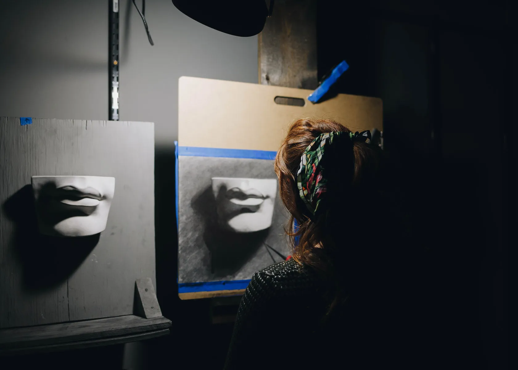 A person with a colorful headscarf is drawing a pair of lips on a gray canvas, illuminated by a focused light. A similar 3D model of lips is mounted to their left for reference. The room appears dimly lit.