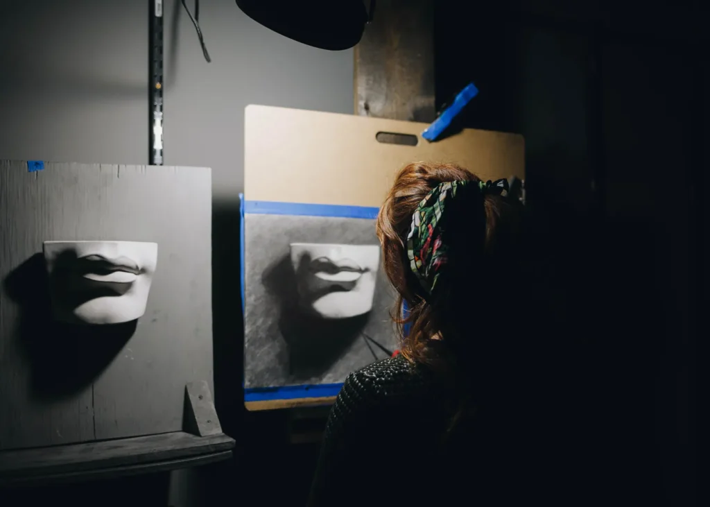 A person stands in a dimly lit art studio, observing a drawing of lips on an easel, with a similar sculpture beside it. The artist's back is turned, and their hair is tied with a patterned scarf. Dramatic shadows enhance the scene's moody ambiance.