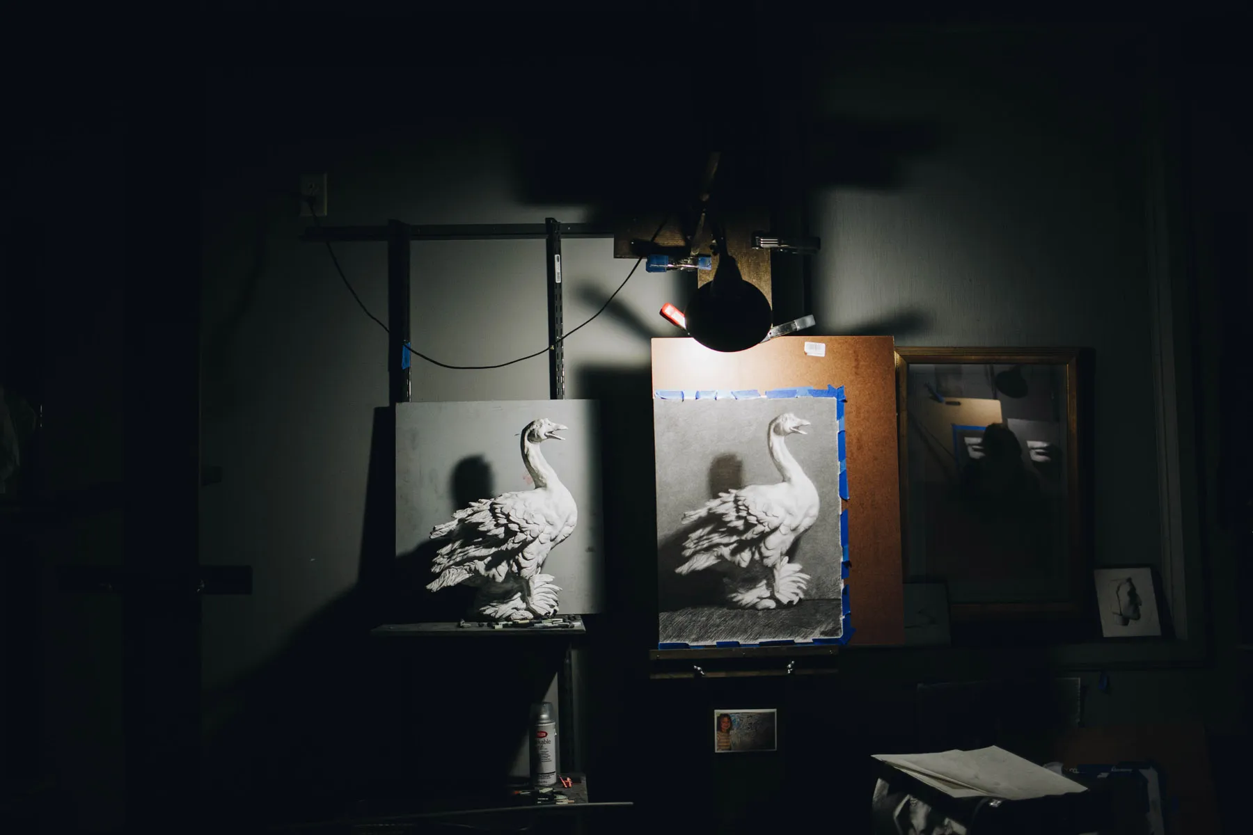 A dimly lit art studio showcases two monochrome swan paintings side by side on easels. A single overhead lamp casts light, creating dramatic shadows. The room is largely dark, emphasizing the artworks.