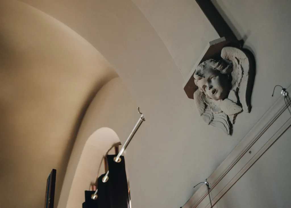 An ornate architectural detail depicting a cherubic face with wings is mounted on a curved, light-colored wall. Below, modern lighting fixtures and wires are visible against the wall. The scene is softly lit, casting gentle shadows.