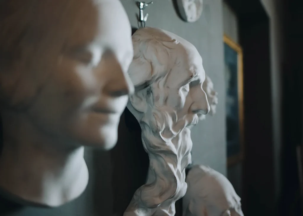 A close-up of two white marble sculptures of human faces in profile view. The foreground sculpture has a smooth, serene expression, while the background sculpture features more intricate details with a textured surface. They are displayed indoors.