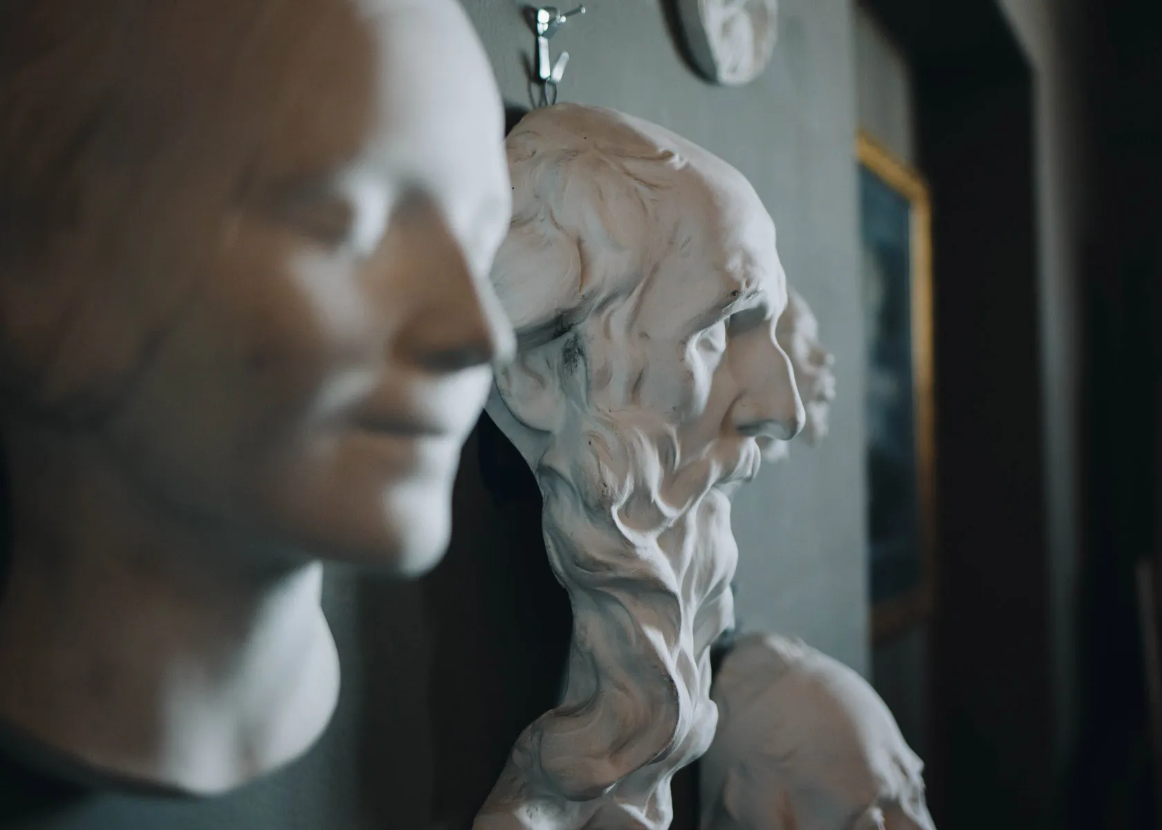 A close-up of two white marble sculptures of human faces in profile view. The foreground sculpture has a smooth, serene expression, while the background sculpture features more intricate details with a textured surface. They are displayed indoors.