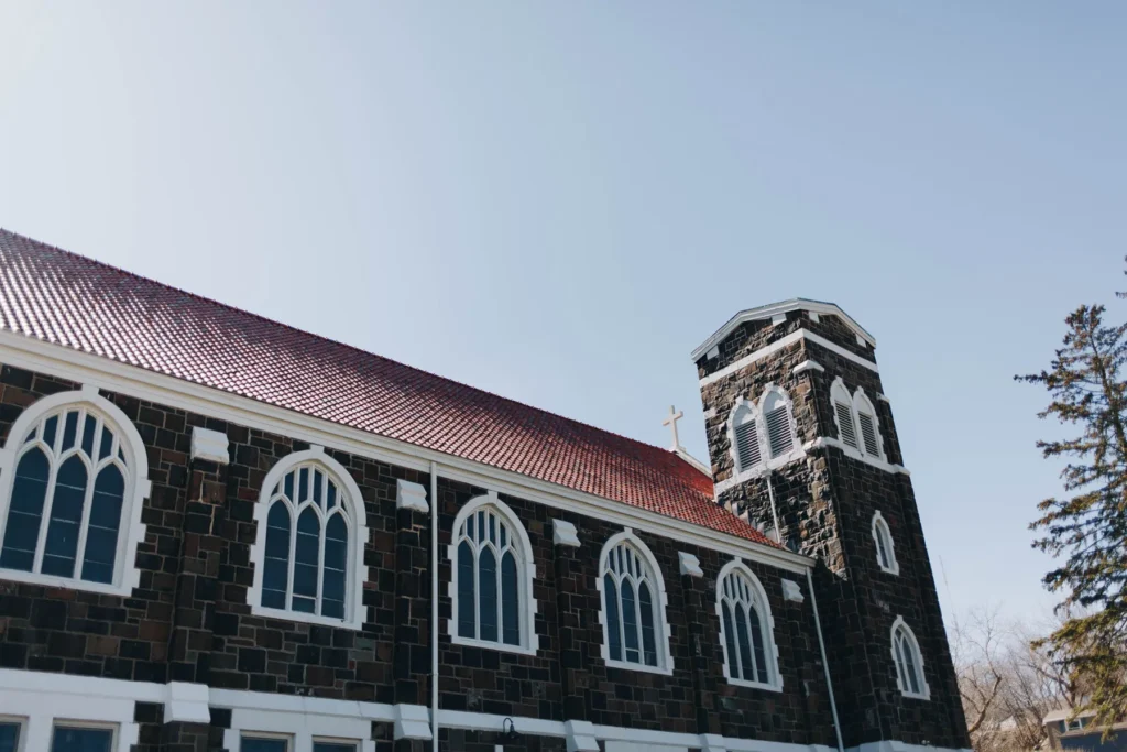 A large stone church with arched windows and a red tiled roof under a clear blue sky. The church features a prominent tower on the right with a cross on top.