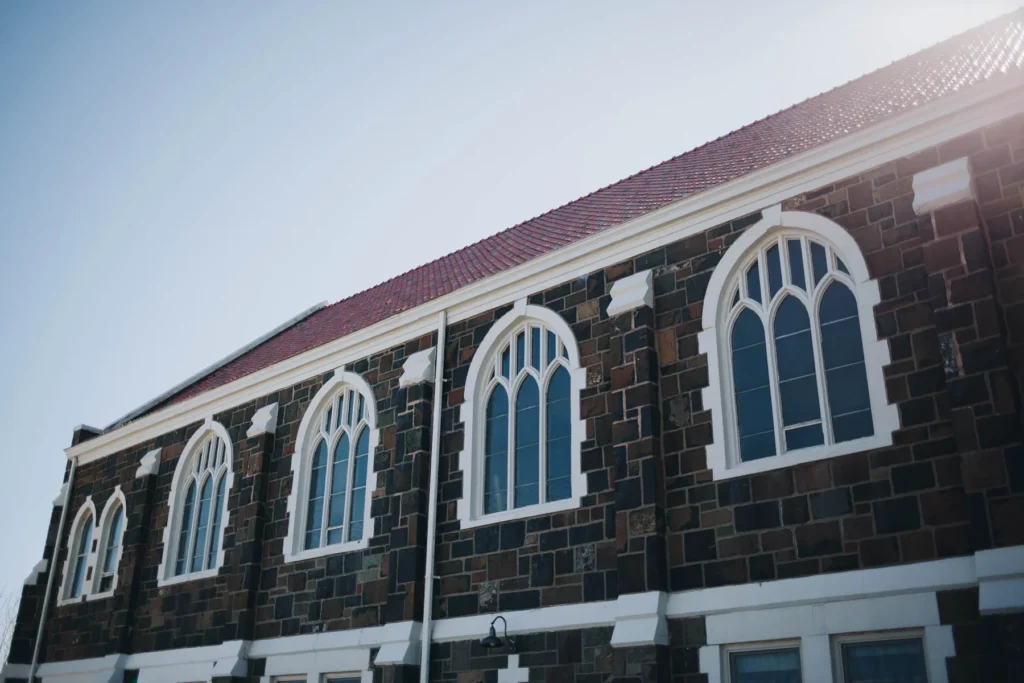 A stone building with large, arched windows and a red tiled roof is seen from below. The windows are divided by white trim. The bright blue sky and sunlight create a serene backdrop.