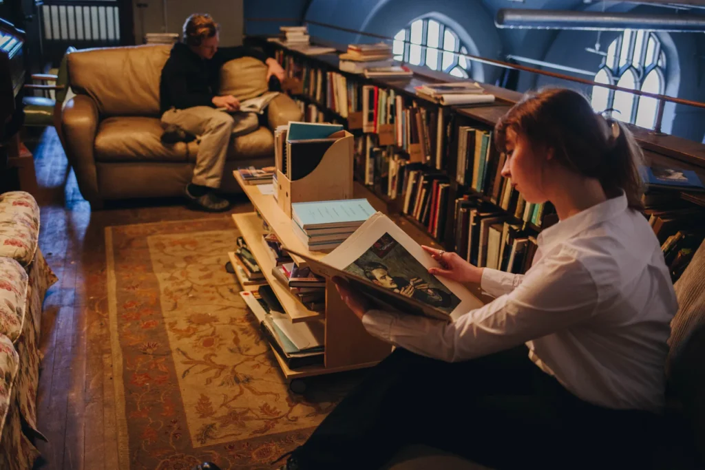 Two people reading in a cozy library. One person sits on a couch with headphones, while the other sits on a bench near a bookshelf, holding an open book. The room has a warm ambiance with books and soft lighting.