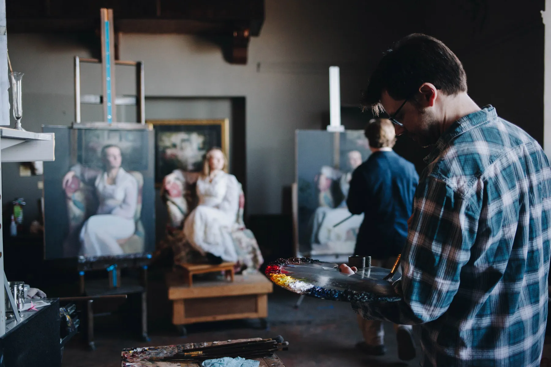 An artist paints a portrait in a dimly lit studio. A woman in white poses on a chair, while another person stands near another canvas. The artist holds a palette with vibrant colors, focused on their work.