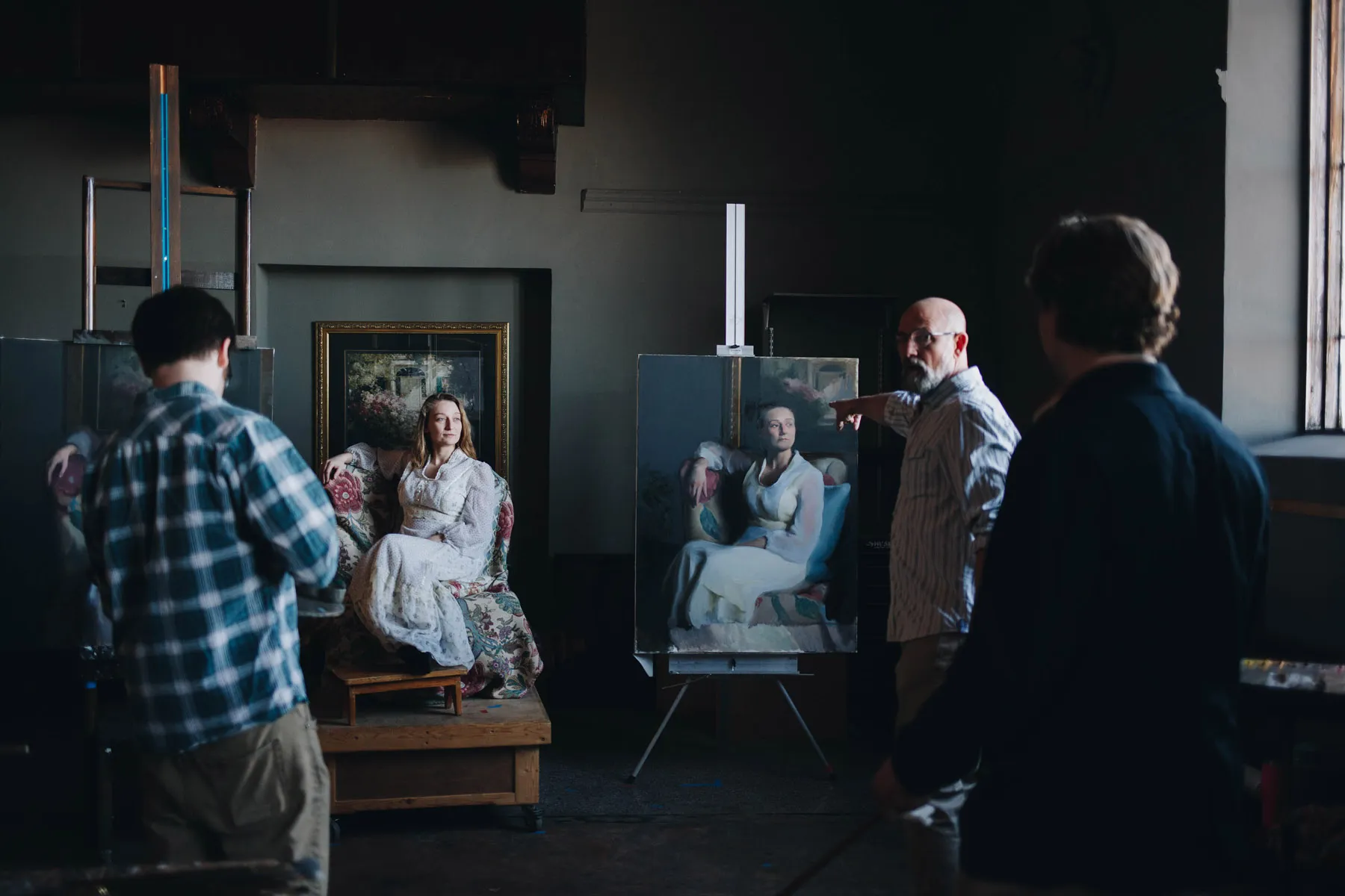 A woman in a white Victorian dress sits as a model in a dimly lit art studio. Artists are positioned around her, painting her portrait. Two completed paintings are visible on easels, capturing her seated pose.