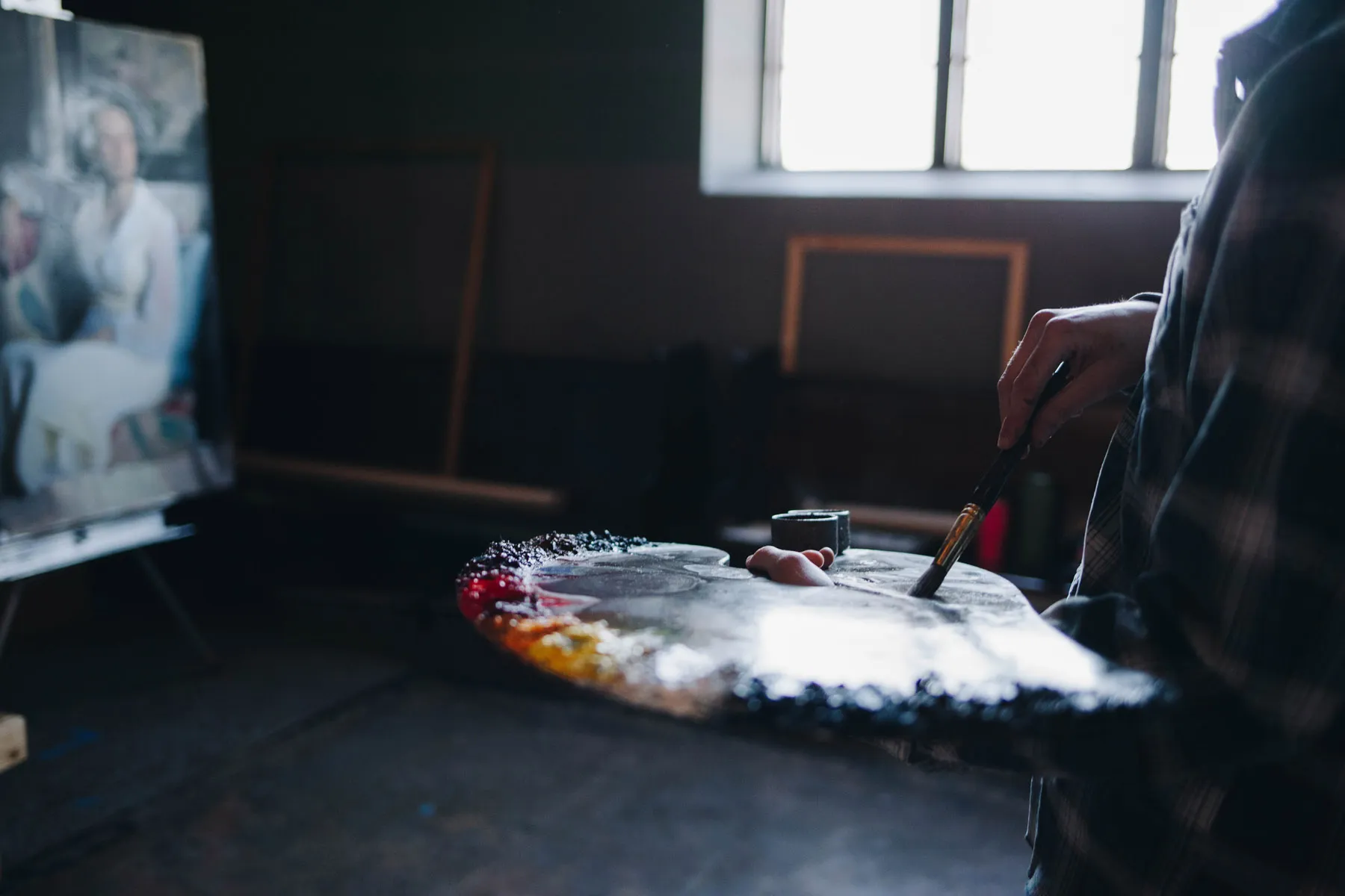 A person holds a paintbrush and palette with various colors in a dimly lit art studio. In the background, there is an easel with a painting near a window letting in light. The focus is on the hand and the palette.